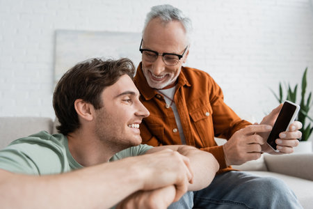 happy and mature father with beard pointing with finger at smartphone near cheerful sonの写真素材