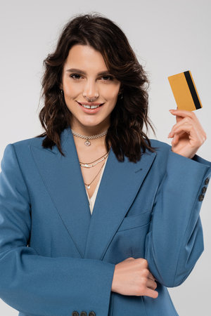cheerful woman in blue blazer and necklaces holding credit card and smiling at camera isolated on...