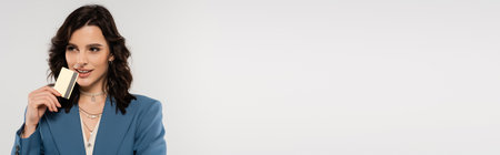 stylish brunette woman in blue jacket looking away while holding credit card isolated on grey, bannerの写真素材