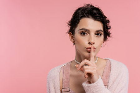 young brunette woman showing hush sign and looking at camera isolated on pink