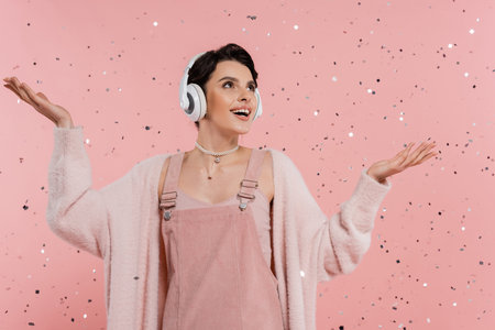 amazed and happy woman in wireless headphones standing under confetti on pink backgroundの写真素材