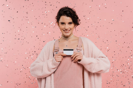 brunette woman in trendy clothes showing credit card and smiling at camera under confetti on pink backgroundの写真素材