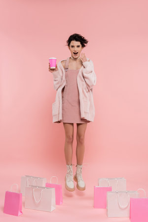 full length of astonished woman with coffee to go levitating near shopping bags on pink backgroundの写真素材