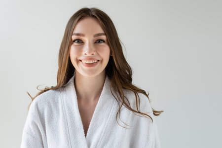 cheerful woman in white bathrobe looking at camera and smiling isolated on greyの写真素材