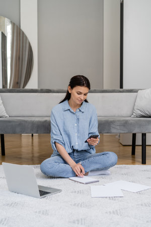 Brunette freelancer using smartphone near notebook and laptop on floor at home
