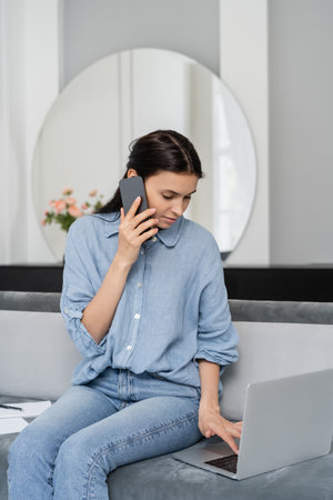 Brunette woman talking on smartphone and using laptop while sitting on couch at homeの写真素材