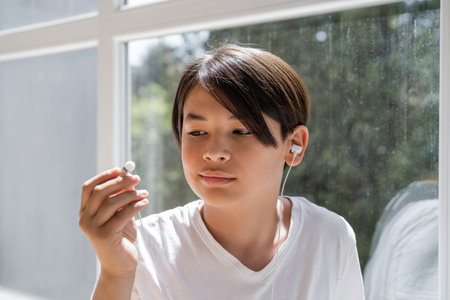 Asian kid looking at wired earphone near window at homeの写真素材