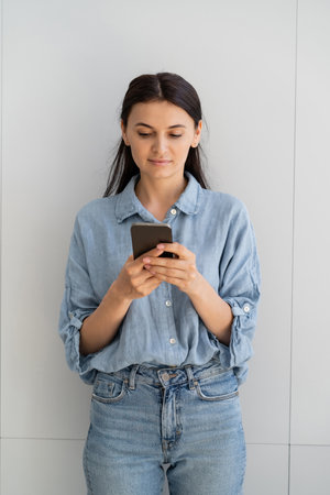 Brunette woman in shirt using smartphone near wall at homeの写真素材