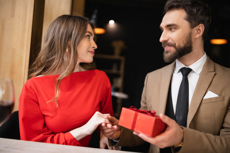 bearded man in formal wear holding present near smiling woman in red dress on valentines dayの写真素材