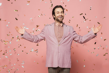 Cheerful host of event pointing at falling confetti and looking at camera on pink backgroundの写真素材