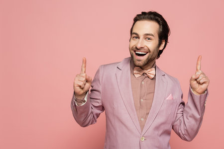 cheerful and young showman in suit with bow tie pointing up with fingers isolated on pinkの写真素材