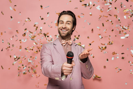 Smiling host of event holding microphone and pointing at camera under confetti on pink backgroundの写真素材