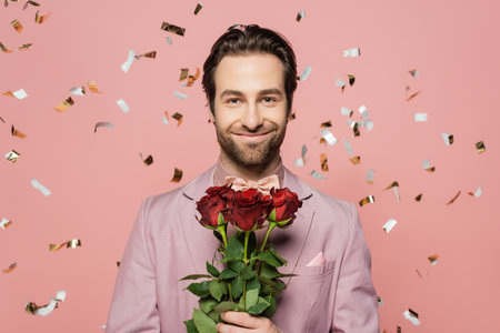 Portrait of positive host of event holding roses under falling confetti on pink backgroundの写真素材