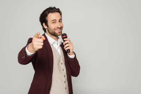 happy showman in formal wear talking in microphone and pointing with finger at camera isolated on greyの写真素材