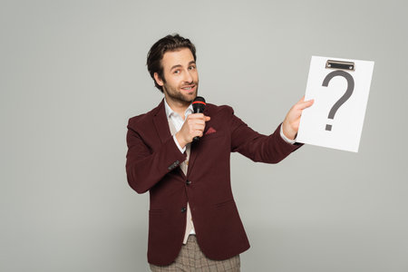 bearded host of event in formal wear talking in microphone while holding clipboard with question...