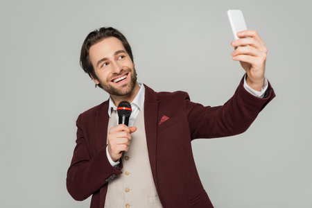 cheerful showman taking selfie on smartphone while holding microphone isolated on greyの写真素材