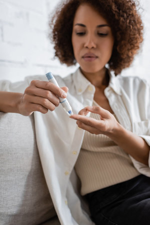 Blurred african american woman with diabetes using lancet pen in living roomの写真素材