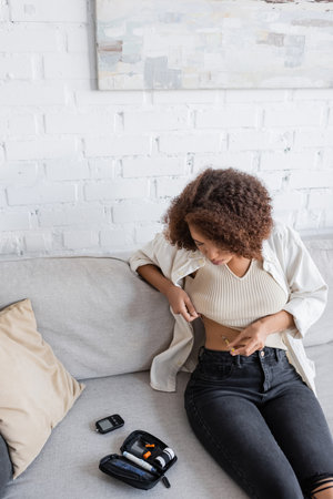 African american woman with diabetes doing insulin injection near medical kit on couchの写真素材