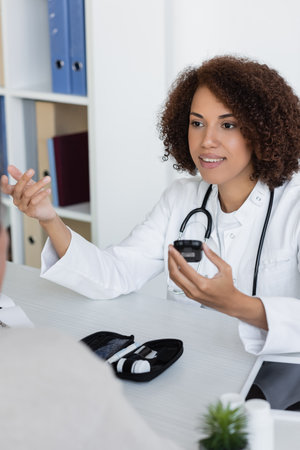 cheerful african american doctor holding glucometer and talking with middle aged patient with...