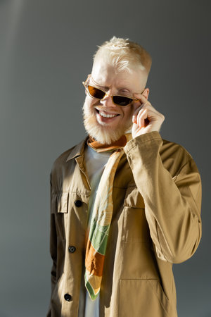 sunlight on face of happy albino man adjusting stylish sunglasses and winking on greyの写真素材