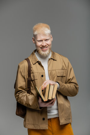 cheerful albino student with backpack holding books isolated on grey