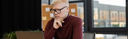 bearded albino businessman in earphones looking at laptop while working in office, bannerの写真素材