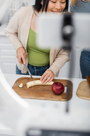 Smiling asian woman cutting banana near friend and blurred smartphone