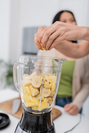 Mature woman squeezing lemon while preparing fruit smoothie with friend in kitchenの写真素材