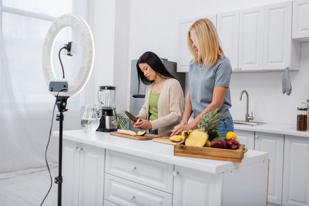 Interracial bloggers cutting fresh fruits near blender and smartphone in kitchen