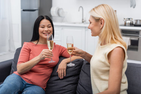 Cheerful asian woman holding champagne and looking at camera near blurred friend at homeの写真素材