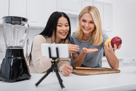 Positive interracial women pointing at fruits near blender and smartphone on tripod in kitchenの写真素材