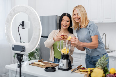 Positive multiethnic women putting fruits in blender near smartphone and ring light in kitchenの写真素材