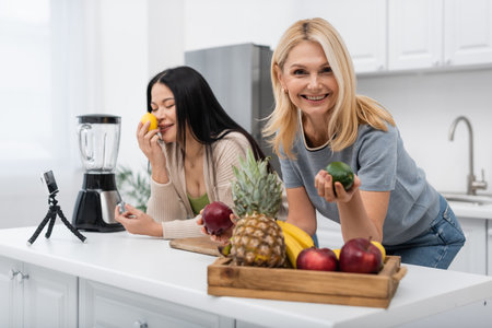 Smiling woman holding fruit near asian friend and smartphone on tripod in kitchenの写真素材
