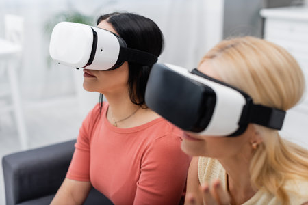 Brunette woman in virtual reality headset gaming near blurred friend at home
