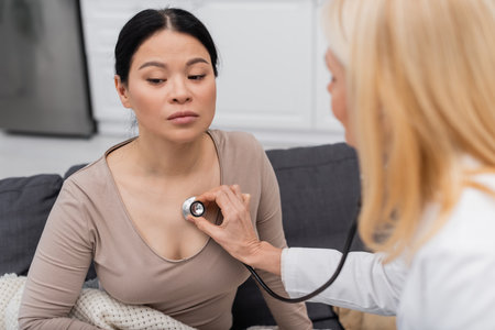 Blurred doctor with stethoscope checking lungs of sick asian woman at homeの写真素材