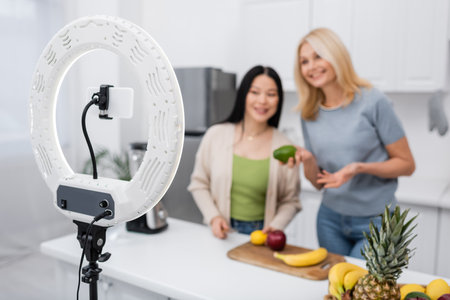 Cellphone and ring lamp near blurred interracial friends with fruits in kitchen