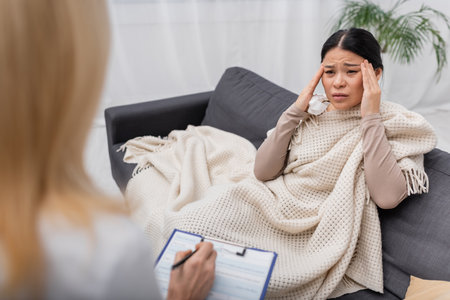 Asian woman holding napkin and suffering from headache near blurred doctor with clipboard at homeの写真素材