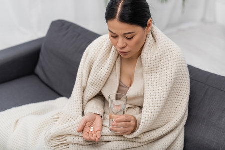 sick asian woman in blanket holding glass of water and pills at homeの写真素材