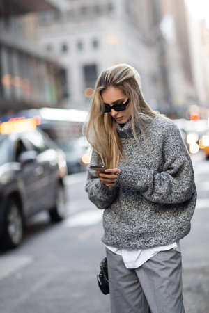concentrated woman in winter sweater and sunglasses using smartphone on urban street in New York cityの写真素材