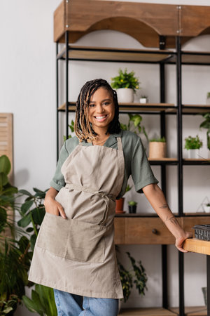 stylish african american woman holding hand in pocket of apron and smiling at camera in flower shopの写真素材