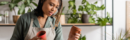 african american florist with braids choosing flowerpots while working in shop, banner