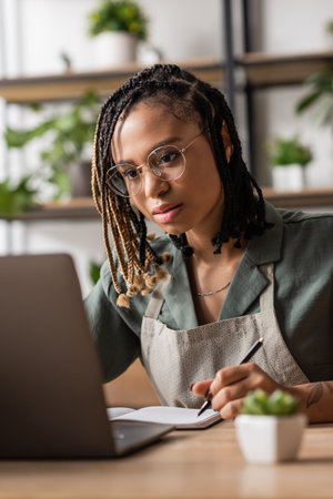 focused african american florist in eyeglasses looking at laptop and writing order in flower shopの写真素材