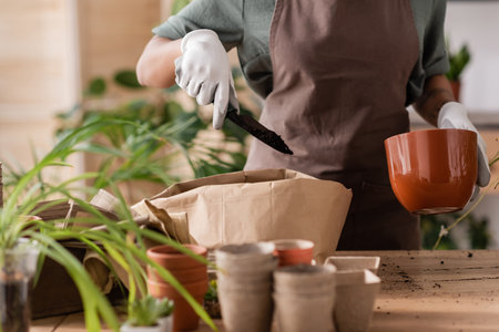 cropped view of african american florist in apron holding flowerpot and garden scoop with soil near...