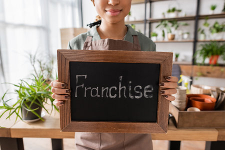 partial view of african american florist holding board with franchise lettering near plants and flowerpots on blurred backgroundの写真素材