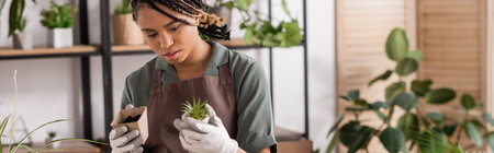 young and stylish african american florist in work gloves holding small green plant and flowerpot with soil in flower shop, bannerの写真素材
