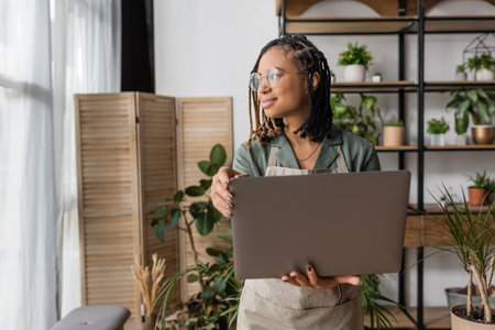 positive african american florist with stylish dreadlocks and eyeglasses holding laptop and looking away in flower shopの写真素材