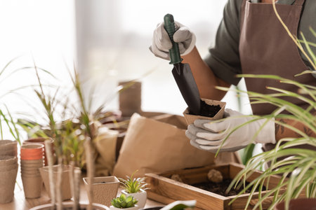 partial view of african american florist holding garden scoop and flowerpot with soil near blurred plantsの写真素材