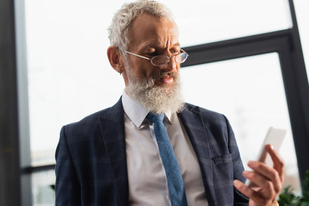 Mature bearded businessman in eyeglasses using blurred smartphone in officeの写真素材