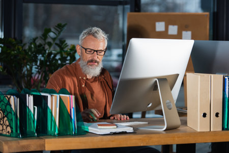Bearded businessman in eyeglasses sitting near computer and paperwork in office in evening