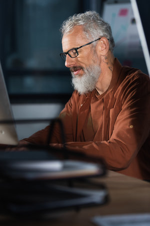 Tattooed businessman in eyeglasses using computer in office in eveningの写真素材